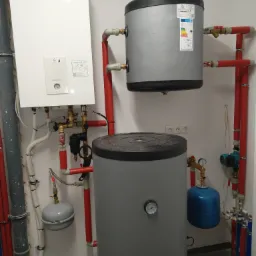 Biały piec gazowy Panasonic zamontowany na ścianie, połączony z dwoma szarymi zbiornikami na wodę, czerwone rury instalacji grzewczej, manometry i zawory w kotłowni.