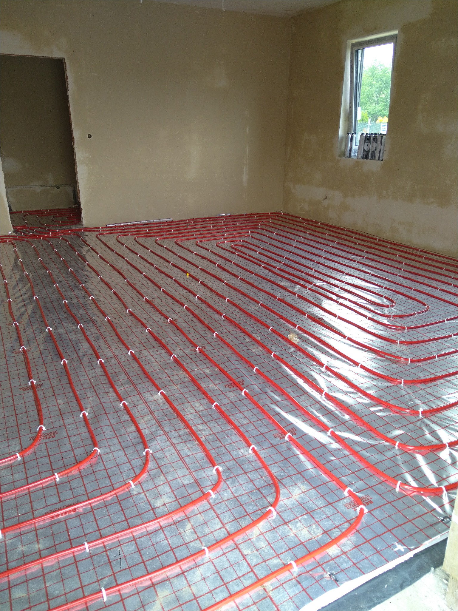 Czerwone rury ogrzewania podłogowego przymocowane do folii z rastrem w surowym wnętrzu z oknem i otwartymi drzwiami.