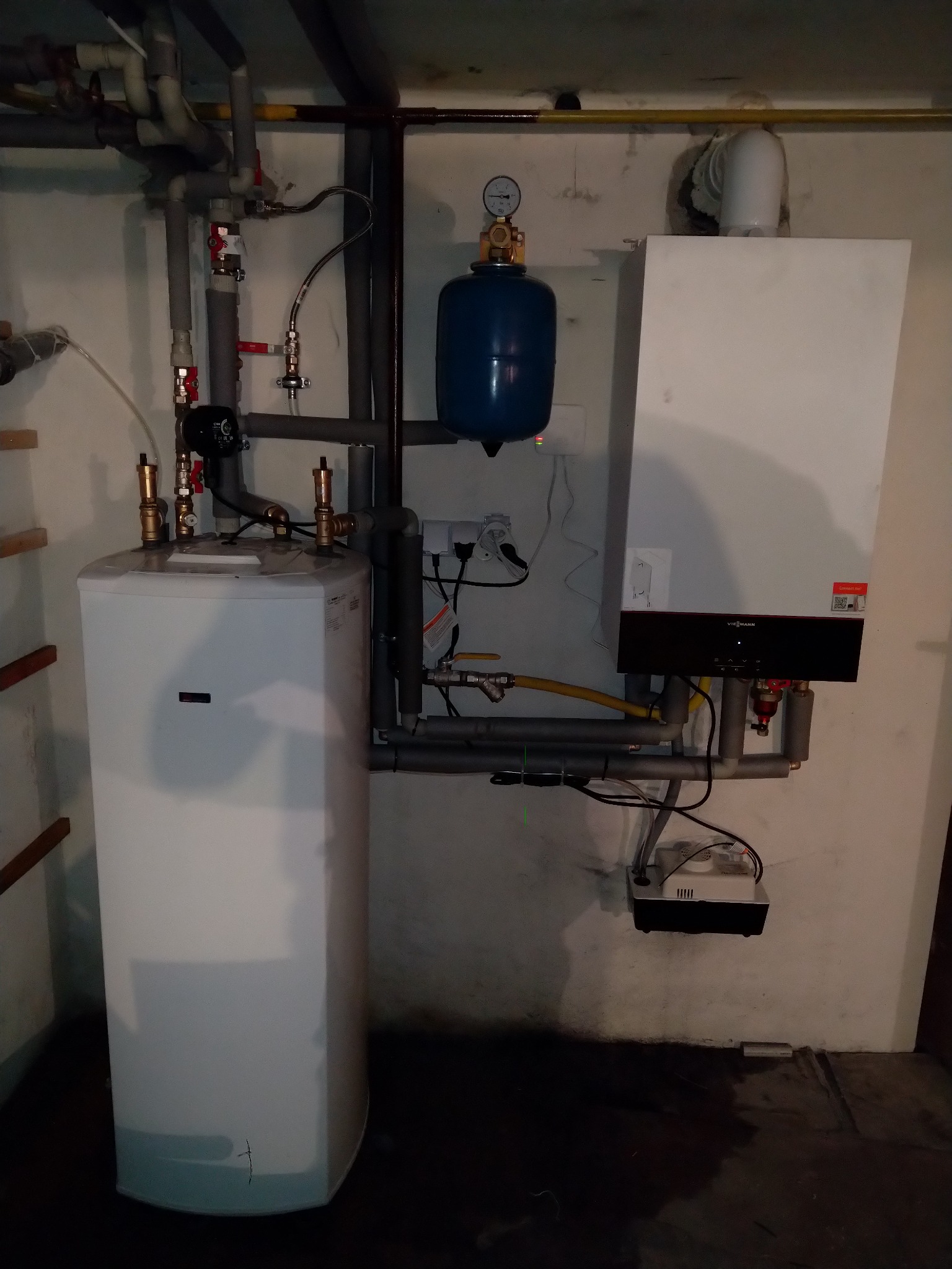 Biały zasobnik ciepłej wody użytkowej połączony z kotłem gazowym Viessmann, widoczne rury instalacyjne, manometr i osprzęt elektryczny, w piwnicy.