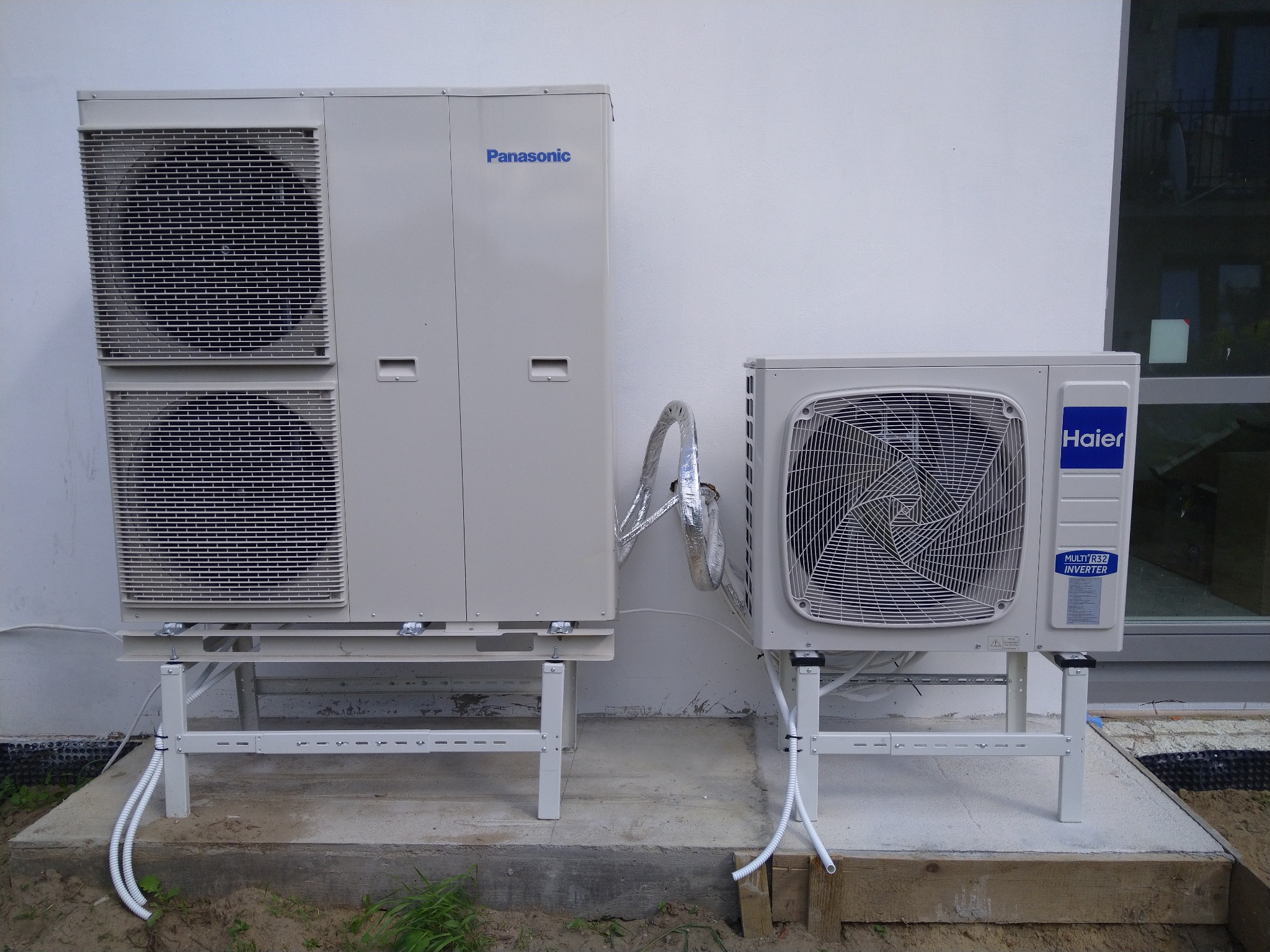 Dwie jednostki zewnętrzne pomp ciepła, Panasonic z podwójnym wentylatorem i Haier, zamontowane na metalowych stelażach na betonowej podstawie przy białej ścianie budynku.