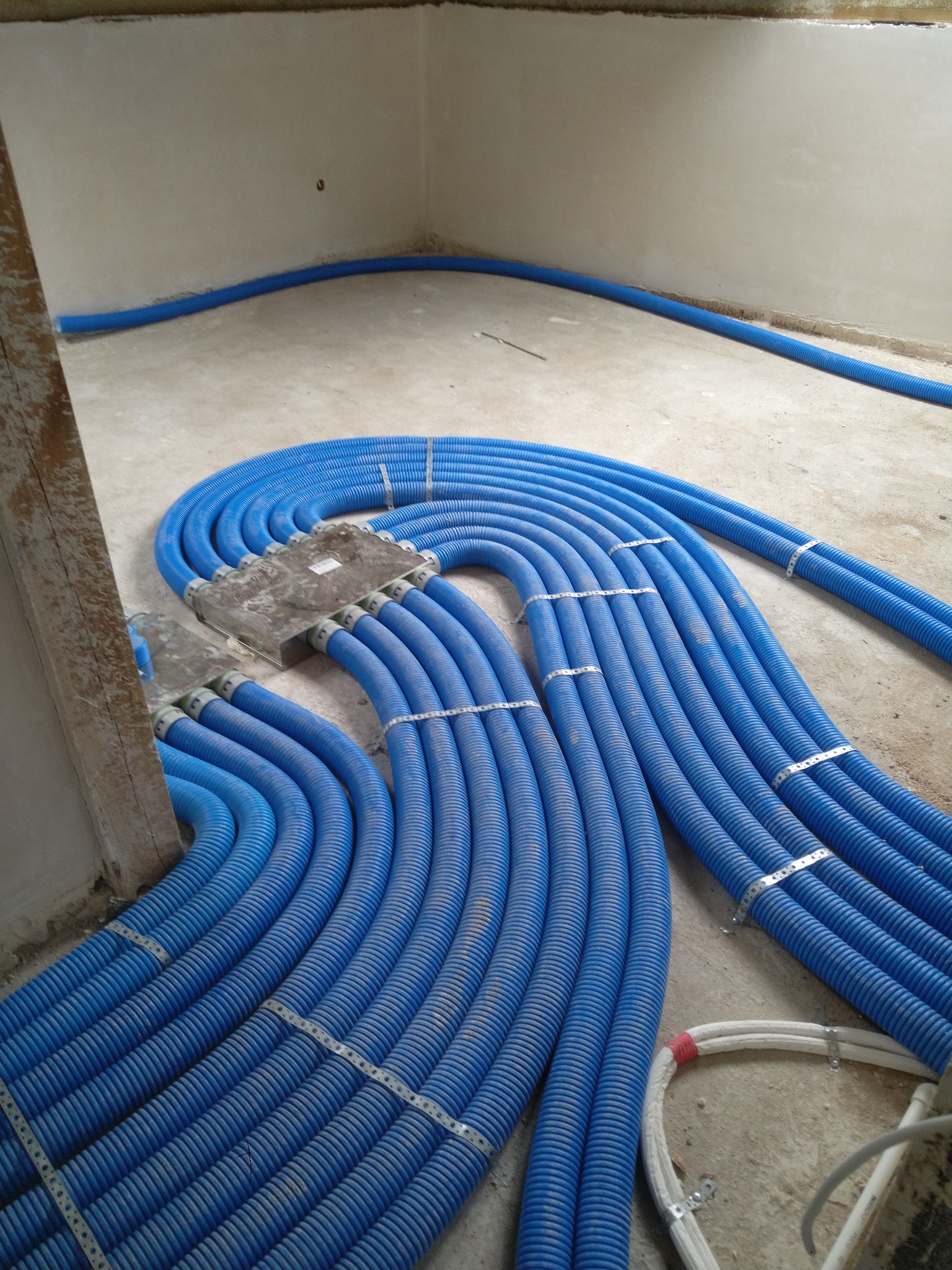 Niebieskie rury karbowane ułożone w pętle na surowym betonowym podłożu, mocowane metalowymi obejmami, widoczne elementy instalacji elektrycznej w tle.