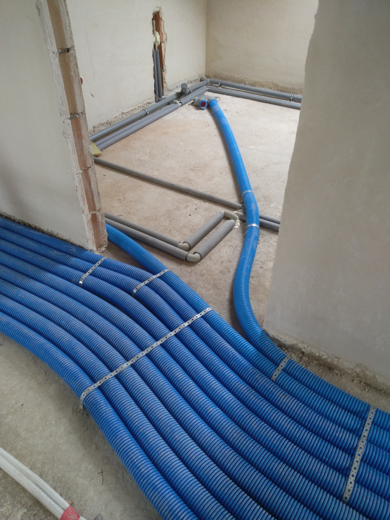 Niebieskie rury karbowane ułożone równolegle, mocowane metalowymi obejmami, podczas montażu instalacji w surowym wnętrzu budynku, widoczne szare rury w tle.