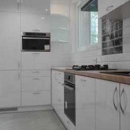 PPUH NIXSTYL - Biała kuchnia z połyskiem, zabudowa z piekarnikiem i szufladami, drewniany blat, płytki imitujące cegłę na ścianie, grafika nad blatem.