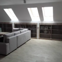 Jasnoszara modułowa sofa z szezlongiem i niskim stolikiem kawowym na tle zabudowy meblowej z przeszklonymi witrynami w pokoju na poddaszu z trzema oknami dachowymi.