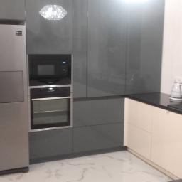 PPUH NIXSTYL - Nowoczesna kuchnia z szarymi, lakierowanymi frontami szafek, srebrną lodówką side-by-side z dozownikiem wody, piekarnikiem i mikrofalówką w zabudowie oraz jasnymi szafkami dolnymi z czarnym blatem.