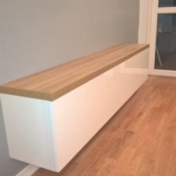 PPUH NIXSTYL - Długa, wisząca szafka z białymi, lakierowanymi frontami i blatem z jasnego drewna, zamontowana na szarej ścianie w pomieszczeniu z parkietem.