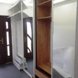 PPUH NIXSTYL - Szafa wnękowa z lustrzanymi drzwiami przesuwnymi, białe półki i szuflady po lewej stronie, drewniane półki i szuflady po prawej, widoczna część wnętrza pomieszczenia i drzwi w odbiciu lustra.