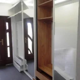 Szafa wnękowa z lustrzanymi drzwiami przesuwnymi, białe półki i szuflady po lewej stronie, drewniane półki i szuflady po prawej, widoczna część wnętrza pomieszczenia i drzwi w odbiciu lustra.