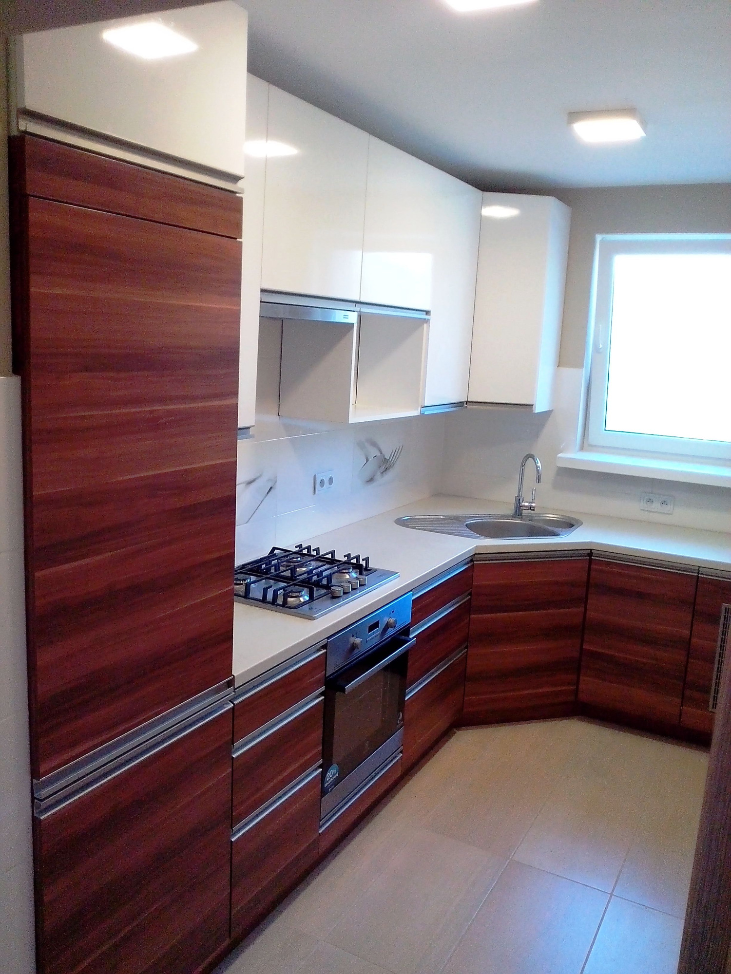 Nowoczesna kuchnia z białymi górnymi szafkami i dolnymi szafkami z ciemnego drewna, srebrne uchwyty, płyta gazowa i piekarnik, jasna podłoga, okno z białymi ramami.