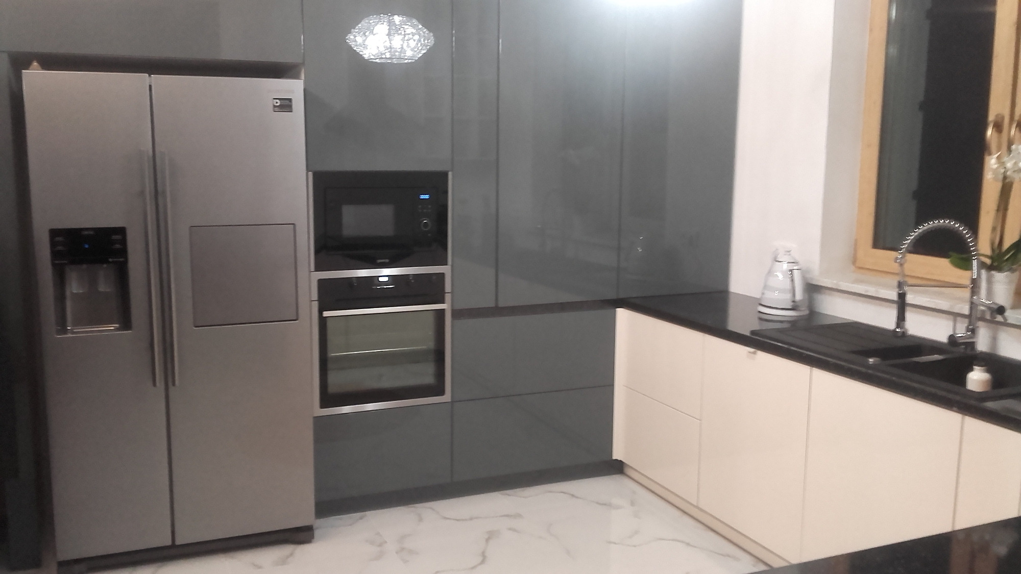 Nowoczesna kuchnia z szarymi, lakierowanymi frontami szafek, srebrną lodówką side-by-side z dozownikiem wody, piekarnikiem i mikrofalówką w zabudowie oraz jasnymi szafkami dolnymi z czarnym blatem.