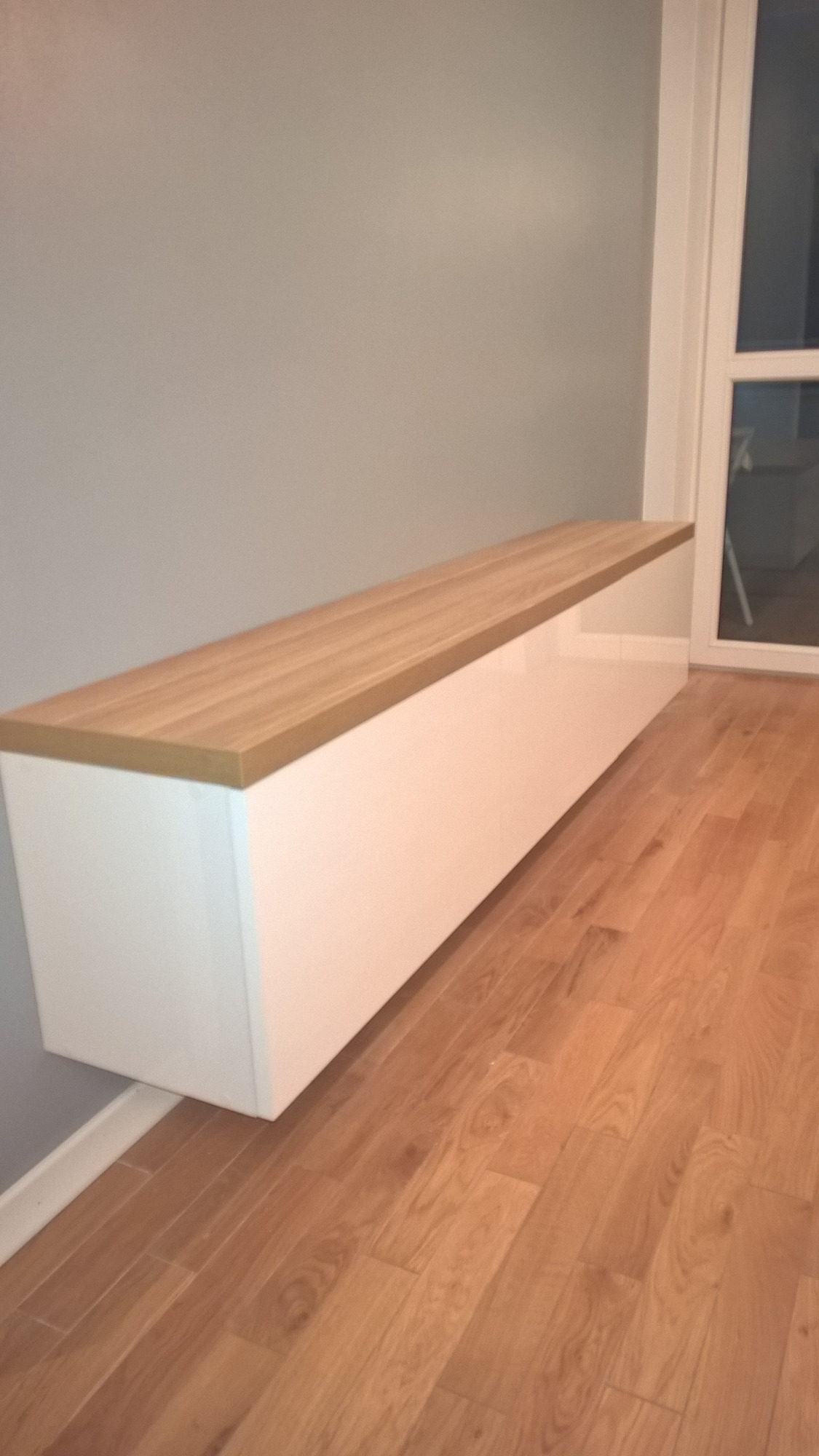 Długa, wisząca szafka z białymi, lakierowanymi frontami i blatem z jasnego drewna, zamontowana na szarej ścianie w pomieszczeniu z parkietem.