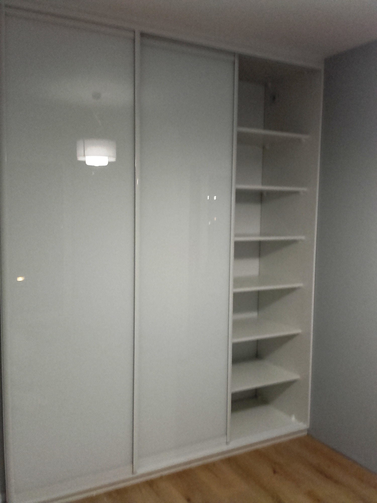Biała szafa wnękowa z przesuwnymi, lustrzanymi drzwiami, częściowo otwarta, ukazująca półki. Widoczna podłoga z paneli.