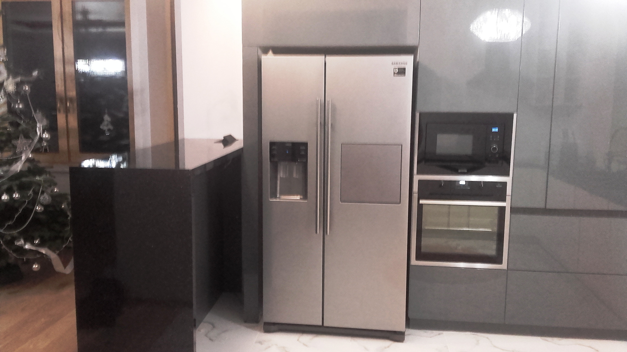 Szara zabudowa kuchenna z dużą lodówką side-by-side, piekarnikiem i mikrofalówką; w tle widoczna choinka.