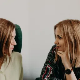 Dwie kobiety siedzą naprzeciwko siebie, prawdopodobnie podczas sesji terapeutycznej. Jedna kobieta w zielonej bluzce siedzi na zielonym krześle, druga w koszuli w paski opiera podbródek na dłoni...