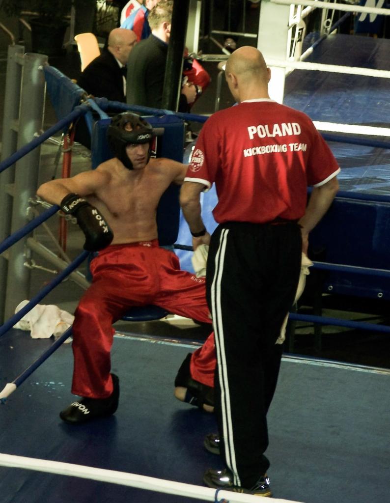 Kickboxer w czerwonym stroju siedzi w rogu ringu, trener w czerwonej koszulce z napisem 'POLAND KICKBOXING TEAM' stoi obok