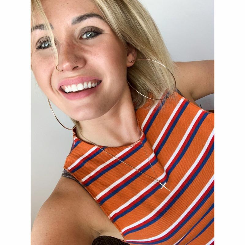 Uśmiechnięta blondynka w pomarańczowej koszulce z paskami, z dużymi złotymi kolczykami kołami i delikatnym naszyjnikiem z krzyżykiem, na jasnym tle.