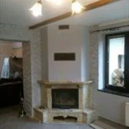 Wykończony kominek narożny z jasnego kamienia w surowym wnętrzu, widoczne niedokończone ściany i fragment podłogi z płytami. W tle okno z widokiem na zewnątrz.