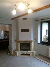 Wykończony kominek narożny z jasnego kamienia w surowym wnętrzu, widoczne niedokończone ściany i fragment podłogi z płytami. W tle okno z widokiem na zewnątrz.