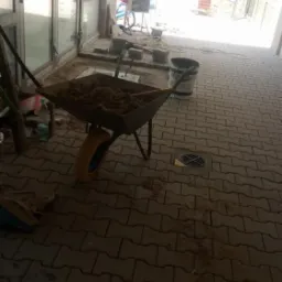 Taczka z piaskiem stoi na ułożonej szarej kostce brukowej w zadaszonym przejściu, w tle widoczne rusztowania i maszyny budowlane.