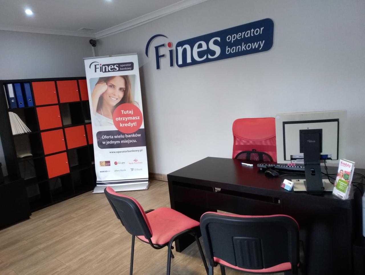 Biuro operatora bankowego z biurkiem, komputerem, krzesłami i roll-upem reklamującym kredyty. Widoczny również regał z segregatorami i logo firmy na ścianie.