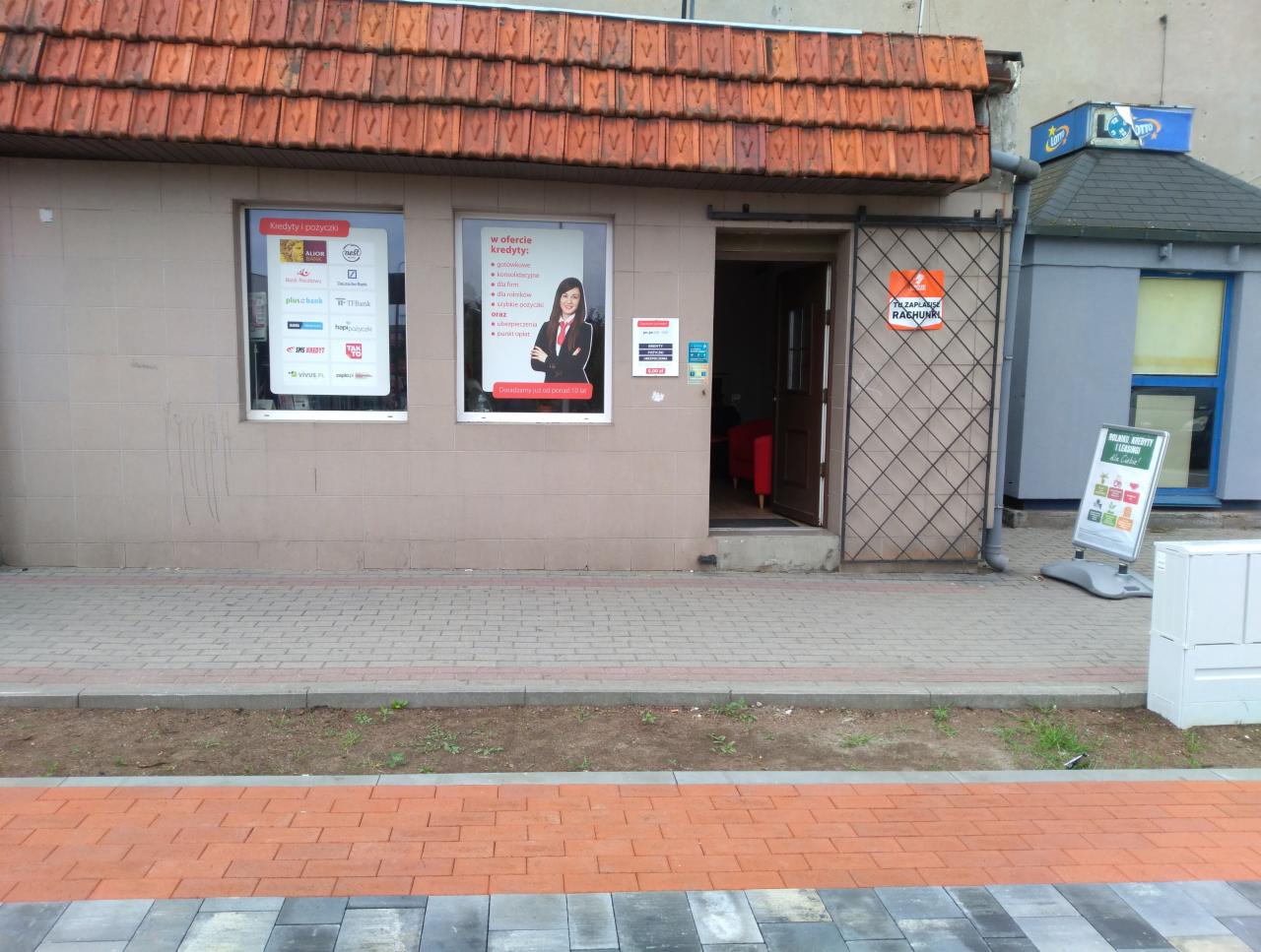 Fasada punktu finansowego z reklamami kredytów i pożyczek w oknach, widoczna tablica informacyjna na zewnątrz, kraty zabezpieczające wejście.
