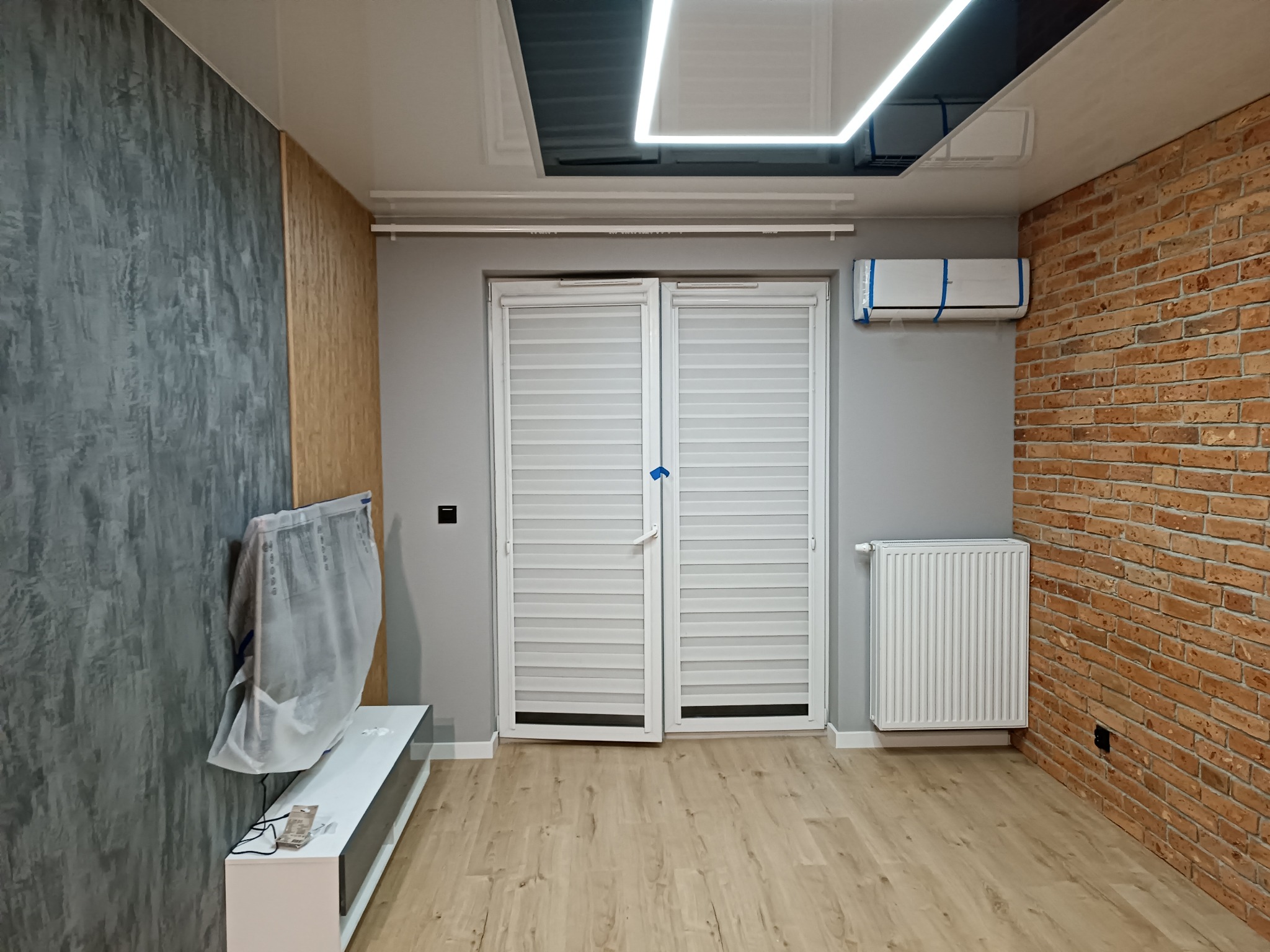 Pokój po remoncie: ściana z cegły, dekoracyjna szara ściana, drewnopodobne panele na podłodze, białe drzwi balkonowe z żaluzjami, grzejnik i klimatyzator zabezpieczony folią.