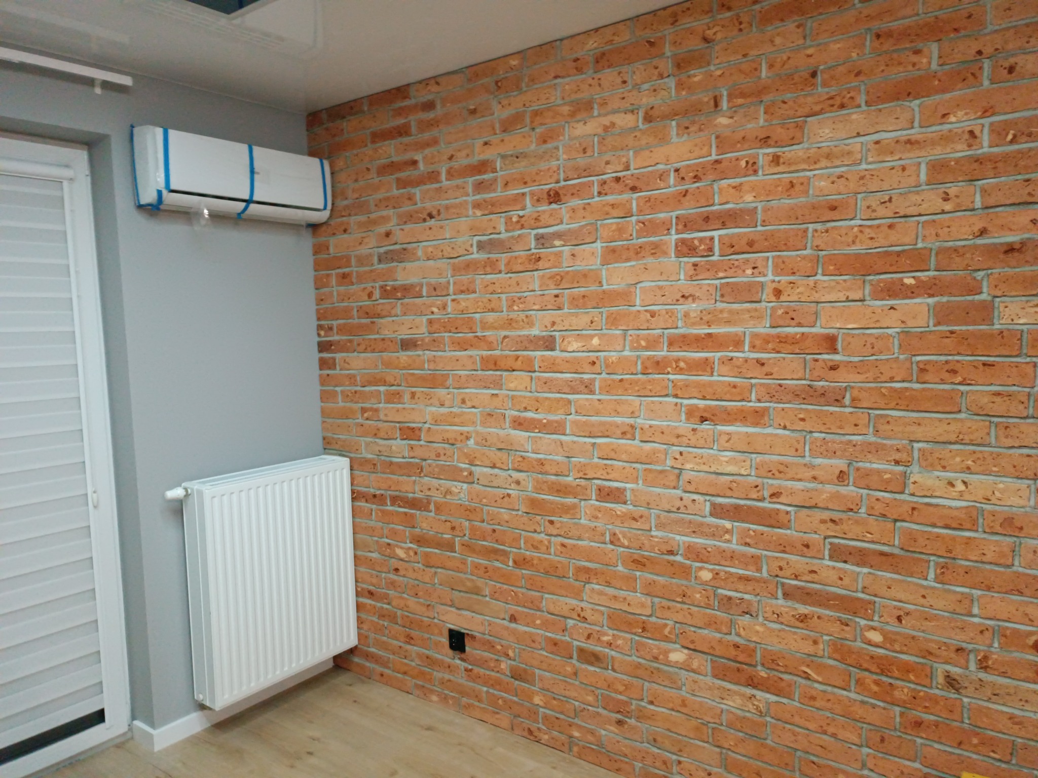 Ściana z cegły dekoracyjnej w stylu loftowym w mieszkaniu w Warszawie, z widocznym grzejnikiem i klimatyzatorem zabezpieczonym folią ochronną.