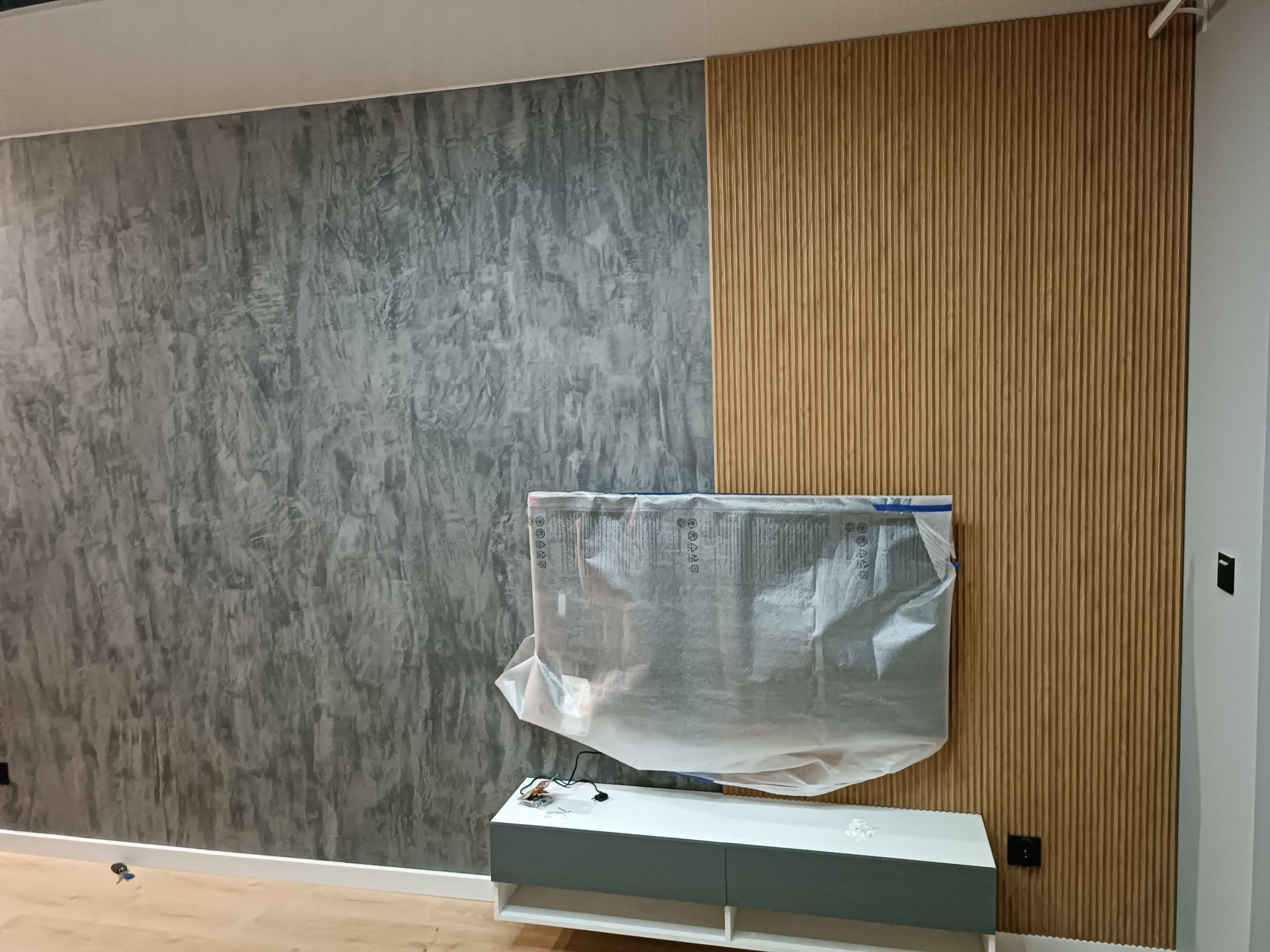 Ściana w pokoju z dekoracyjną szarą strukturą imitującą beton oraz panelami ściennymi z pionowych drewnianych listewek. Telewizor zabezpieczony folią ochronną na szafce RTV.