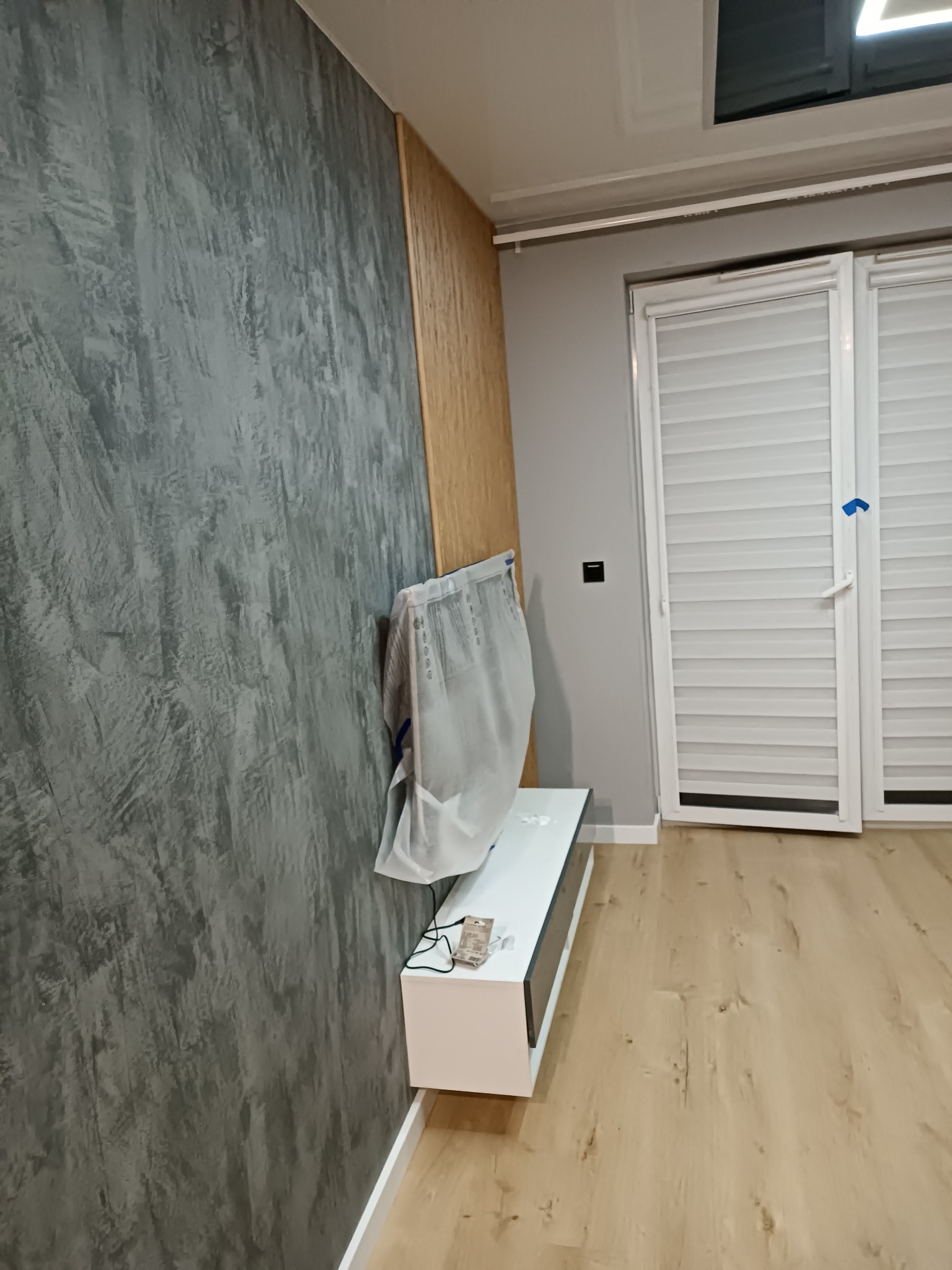 Ściana z efektem dekoracyjnym przypominającym beton, częściowo zakryta folią ochronną, chroniącą meble podczas prac wykończeniowych, nowoczesne wnętrze z drewnianą podłogą i białymi roletami.