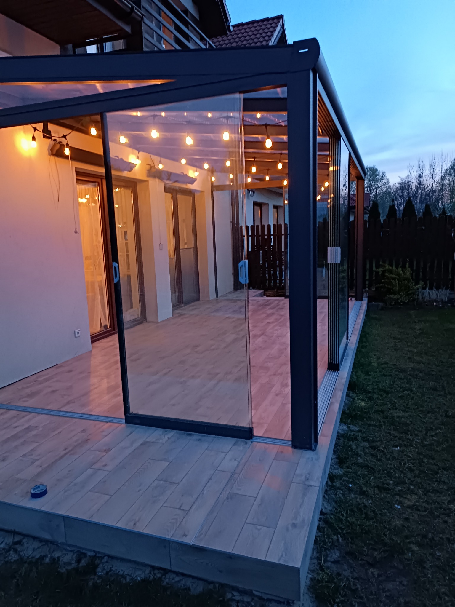 Zadaszony taras z przesuwnymi szklanymi ścianami, oświetlony girlandą żarówek, wykończony płytkami imitującymi drewno, widok z zewnątrz o zmierzchu.