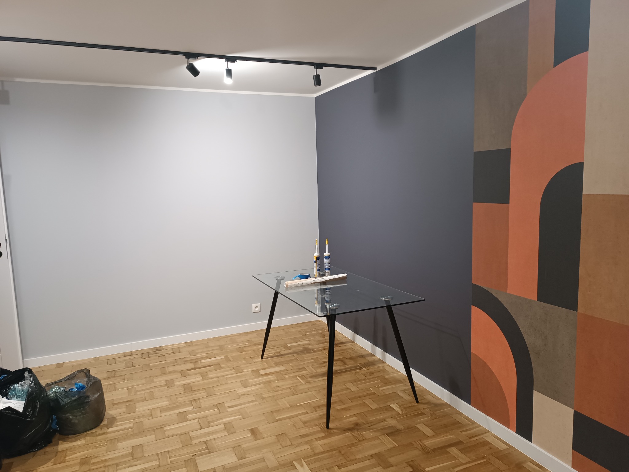 Przygotowania do prac wykończeniowych: stół ze szklanym blatem, na nim tuba z klejem i miarka, ściana częściowo pomalowana na szaro, a częściowo pokryta tapetą w geometryczne wzory, na podłodze...