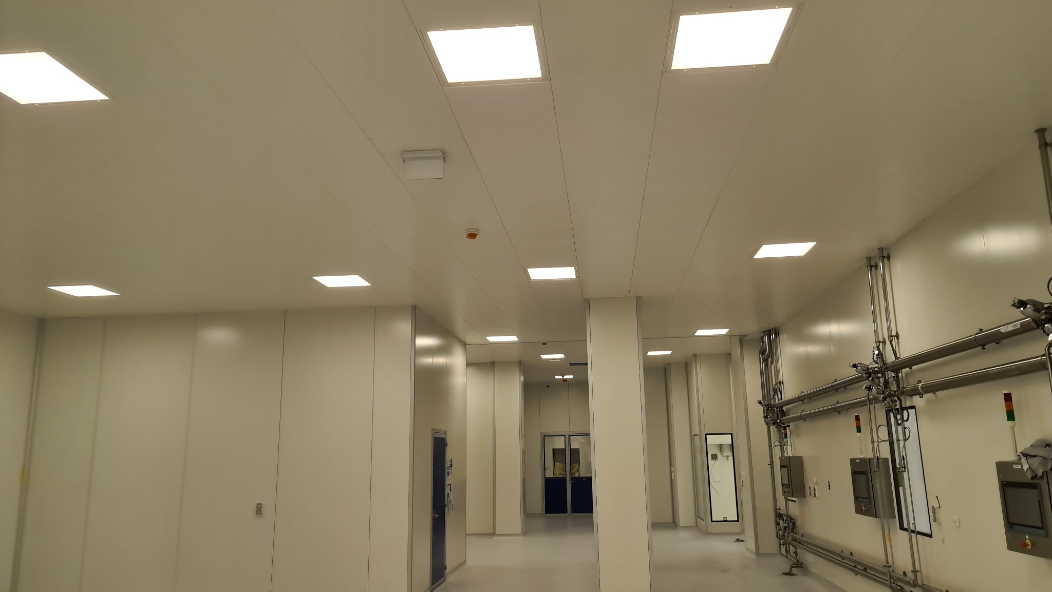 Wnętrze sterylnego pomieszczenia z białymi panelami ściennymi i sufitowymi, kwadratowymi lampami wpuszczanymi, systemem rur i urządzeń kontrolnych na ścianie.