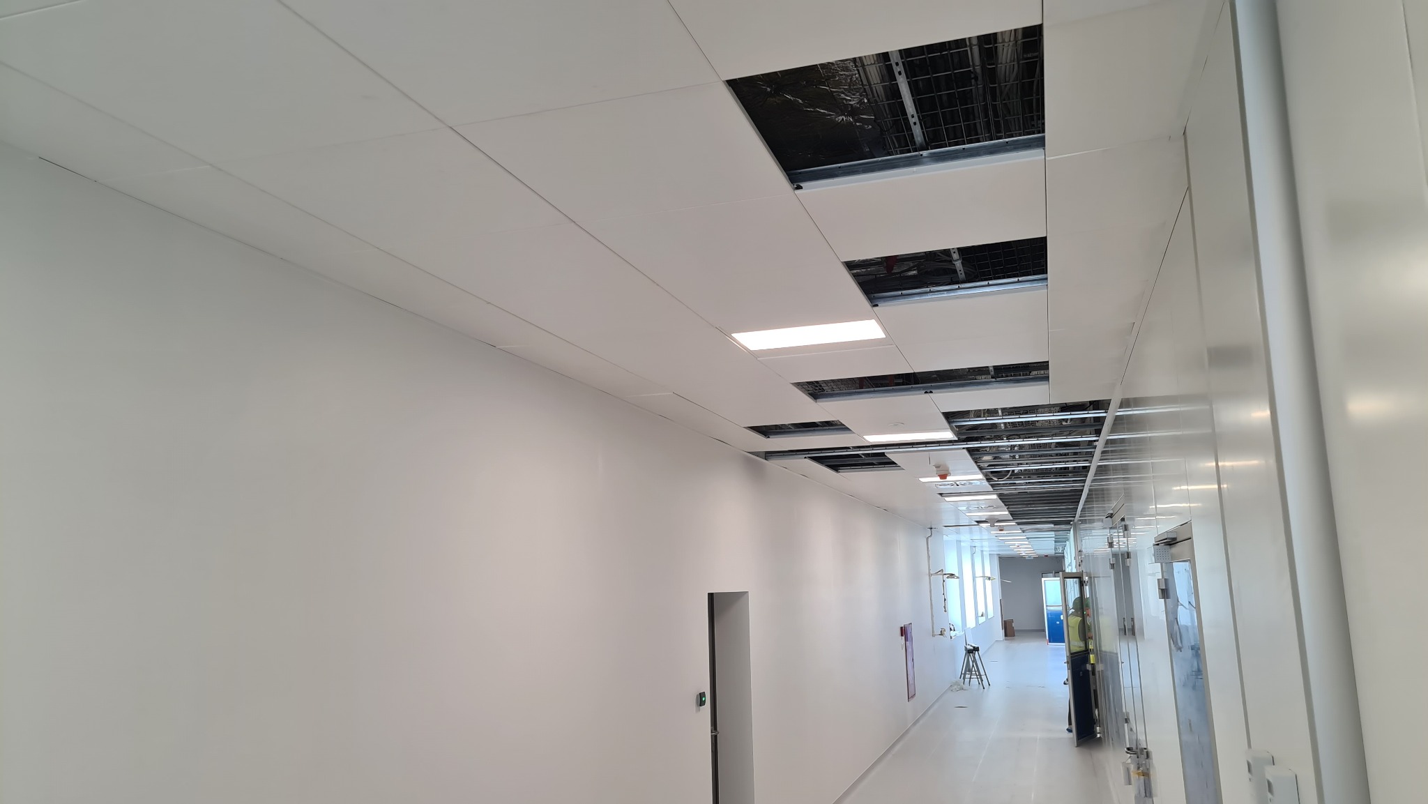 Korytarz biurowy w trakcie montażu sufitu podwieszanego kasetonowego z widoczną instalacją nad sufitem i pracownikiem w oddali.