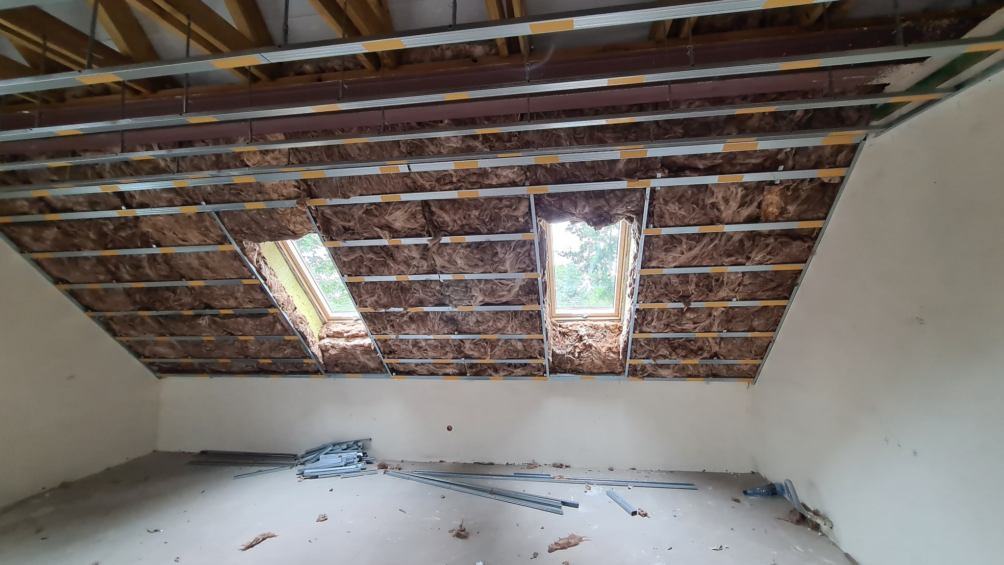 Montaż izolacji z wełny mineralnej w konstrukcji sufitu podwieszanego z dwoma oknami dachowymi; widoczne profile metalowe i pozostałości materiałów na podłodze.