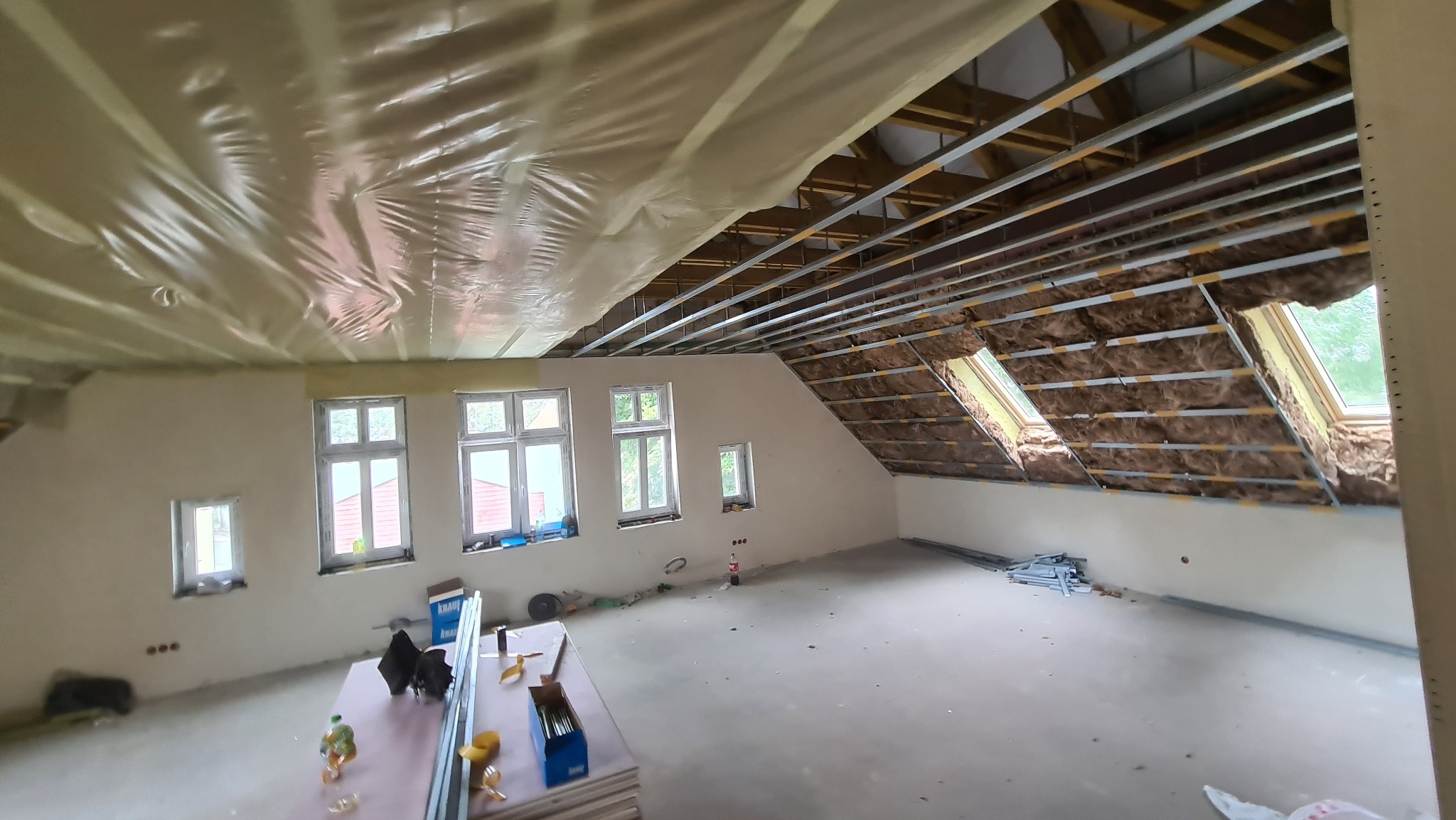 Poddasze w trakcie adaptacji: widoczne metalowe profile sufitowe, izolacja wełną mineralną na skosach, folia paroizolacyjna na suficie, okna dachowe i pionowe. Na podłodze leżą płyty i narzędzia.