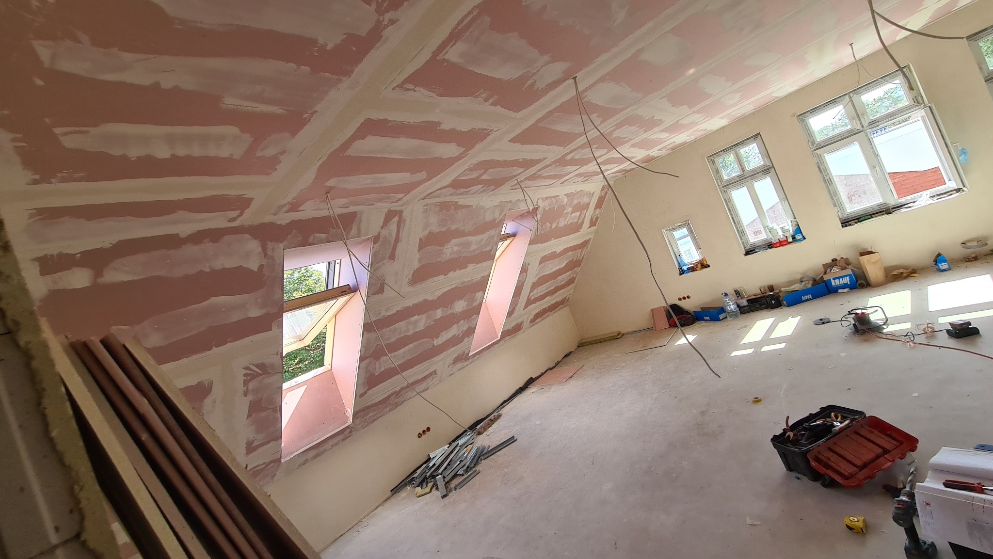 Poddasze w trakcie prac wykończeniowych, widoczne płyty gipsowo-kartonowe na skosach sufitu i ścian, zaszpachlowane łączenia, trzy okna dachowe oraz okna w ścianie szczytowej, skrzynka...