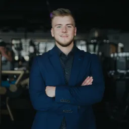 Portret młodego mężczyzny w niebieskim garniturze, stojącego na tle sali z sprzętem do ćwiczeń.