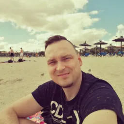 Mężczyzna na plaży z parasolami w tle, uśmiechnięty, w ciemnej koszulce z białym nadrukiem. W tle widać spacerujących ludzi i budynki.