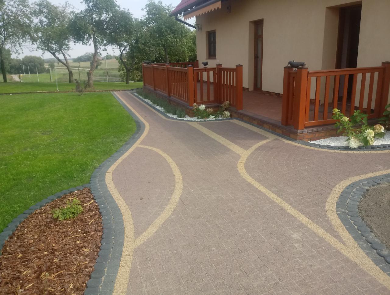 Ułożona kostka brukowa w odcieniach brązu i beżu, z dekoracyjnymi obrzeżami i elementami z kory drzewnej, prowadząca do tarasu domu z drewnianą balustradą.