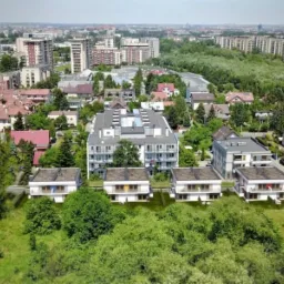 Widok z lotu ptaka na kompleks mieszkalny składający się z nowoczesnego apartamentowca i szeregu domów jednorodzinnych z płaskimi dachami, otoczonych zielenią i zabudową miejską w tle.