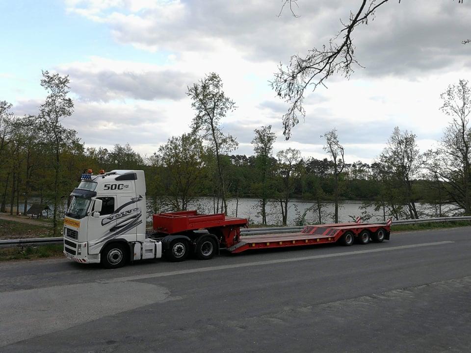 Biała ciężarówka Volvo z niskopodwoziową czerwoną naczepą, zaparkowana na asfaltowej drodze w pobliżu jeziora i drzew, pochmurne niebo w tle.