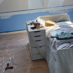 Rafal Gładysz - Pokój na poddaszu w trakcie malowania, zabezpieczony folią ochronną, widoczne narzędzia malarskie, okna dachowe i fragment pomalowanej na niebiesko ściany.