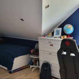 Rafal Gładysz - Pokój dziecięcy na poddaszu z niebieskimi ścianami, białymi meblami, biurkiem z pluszakami na parapecie i łóżkiem z pluszakami, oświetlony światłem dziennym.