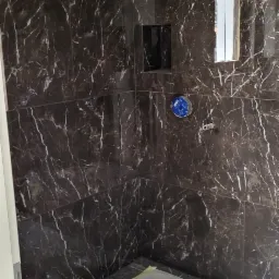 Ciemna, marmurkowa okładzina prysznicowa z wnękami na kosmetyki i niebieskim elementem armatury, zabezpieczona folią podłoga w trakcie prac.