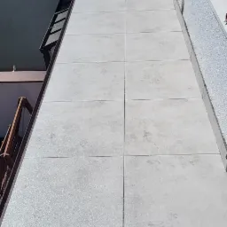 Wyłożony jasnoszarymi płytkami balkon z widocznymi fugami, fragment dachu budynku i buta w prawym dolnym rogu kadru.