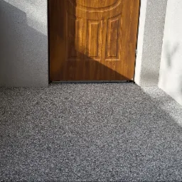 Szara, żywiczna posadzka na zewnątrz budynku, z widoczną drewnianą stolarką drzwiową i fragmentem ściany o szorstkiej fakturze, oświetlona promieniami słońca rzucającymi cień drzewa.