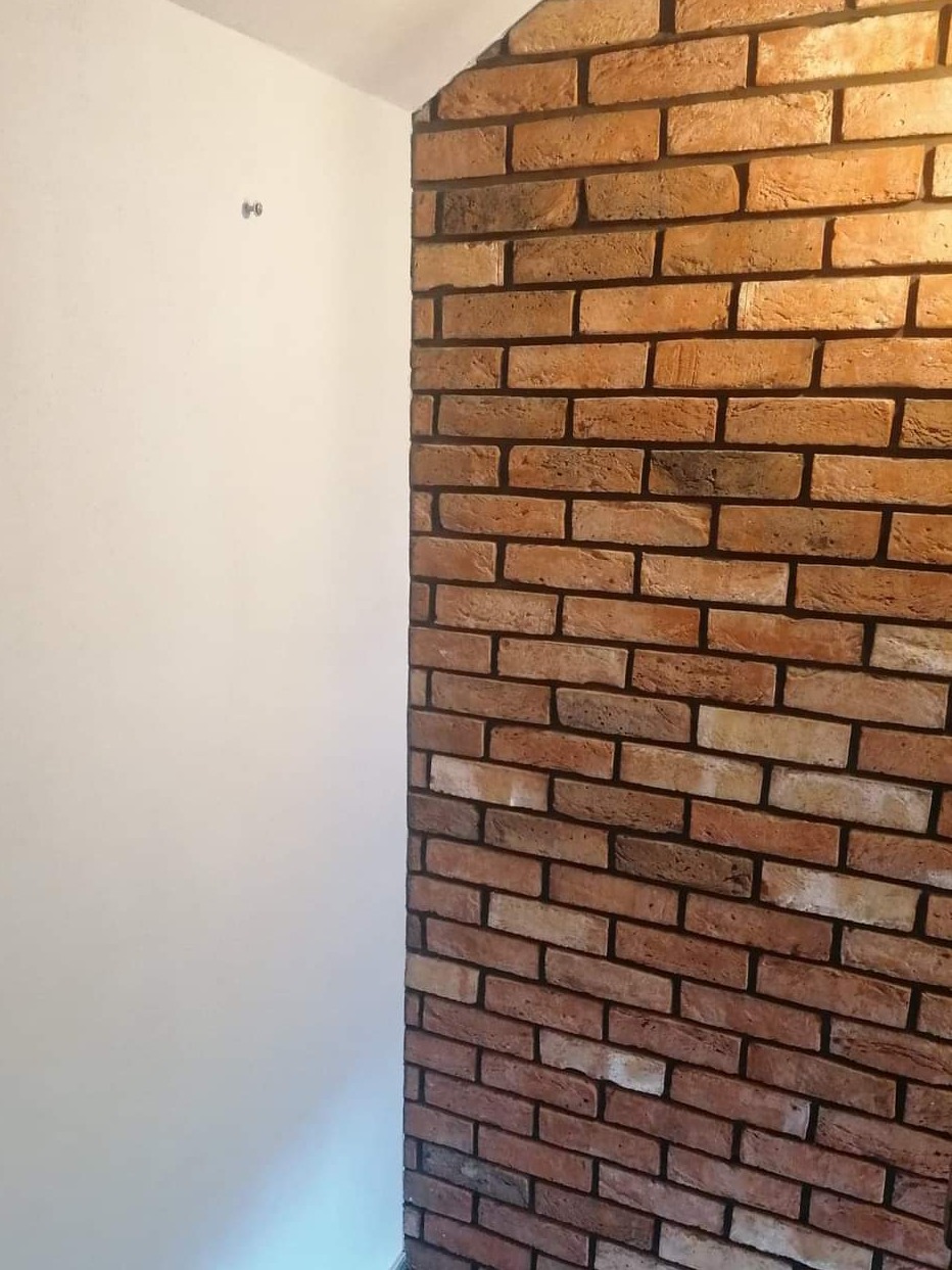 Fragment ściany z cegieł dekoracyjnych w odcieniach brązu i beżu, przylegający do gładkiej, białej ściany w rogu pomieszczenia.