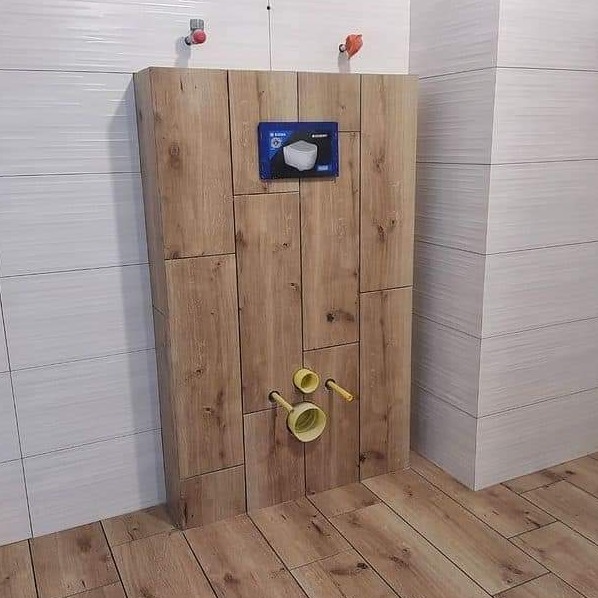 Stelaż podtynkowy WC obudowany drewnopodobnymi panelami z widocznymi przyłączami hydraulicznymi i przyciskiem spłukującym na tle białych płytek ściennych i podłogi z paneli drewnopodobnych.