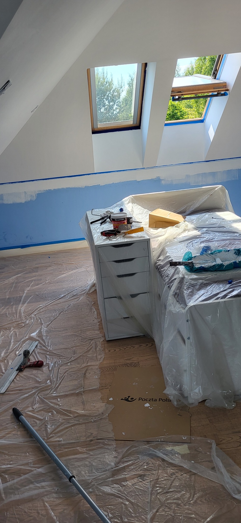 Pokój na poddaszu w trakcie malowania, zabezpieczony folią ochronną, widoczne narzędzia malarskie, okna dachowe i fragment pomalowanej na niebiesko ściany.