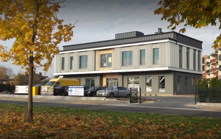 Dwupiętrowy budynek biurowy z szarymi panelami i napisem 'Studio 65' nad wejściem, widoczny z zewnątrz w jesiennej scenerii z drzewem o żółtych liściach i zaparkowanymi samochodami.